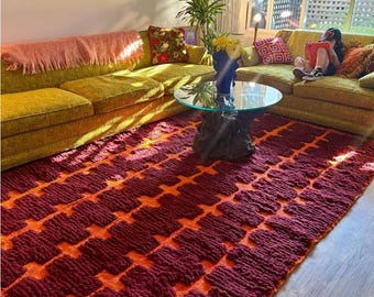 May include: A living room with a burgundy and orange patterned rug. The rug's geometric design dominates the space. Two olive green sofas, a glass-topped coffee table, and decorative cushions create a retro atmosphere. The room is bathed in natural light.