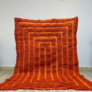 K&ouml;nnte beinhalten: Ein orangefarbener und rostfarbener Teppich mit geometrischem Design. Der Teppich weist eine Reihe vertikaler Linien und ein quadratisches Muster in der Mitte auf. Der Teppich hat ein strukturiertes, weiches Aussehen und einen wei&szlig;en Fransen am unteren Rand.