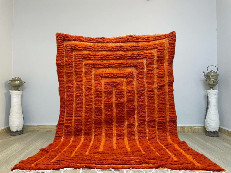 K&ouml;nnte beinhalten: Ein orangefarbener und rostfarbener Teppich mit geometrischem Design. Der Teppich weist eine Reihe vertikaler Linien und ein quadratisches Muster in der Mitte auf. Der Teppich hat ein strukturiertes, weiches Aussehen und einen wei&szlig;en Fransen am unteren Rand.