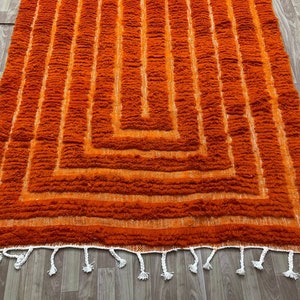 K&ouml;nnte beinhalten: Ein orangefarbener Teppich mit geometrischem Muster. Der Teppich hat eine zottelige Textur mit vertikalen Linien und einem quadratischen Design. Wei&szlig;e Quasten s&auml;umen den unteren Rand.