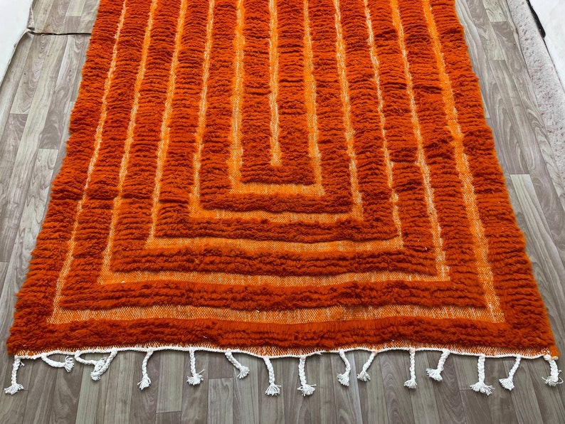 K&ouml;nnte beinhalten: Ein orangefarbener Teppich mit geometrischem Muster. Der Teppich hat eine zottelige Textur mit vertikalen Linien und einem quadratischen Design. Wei&szlig;e Quasten s&auml;umen den unteren Rand.