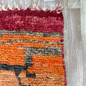 Puede incluir: Una alfombra marroqu&iacute; colorida con un patr&oacute;n geom&eacute;trico en tonos rojos, naranjas y negros. La alfombra tiene un borde de flecos y est&aacute; hecha de lana.
