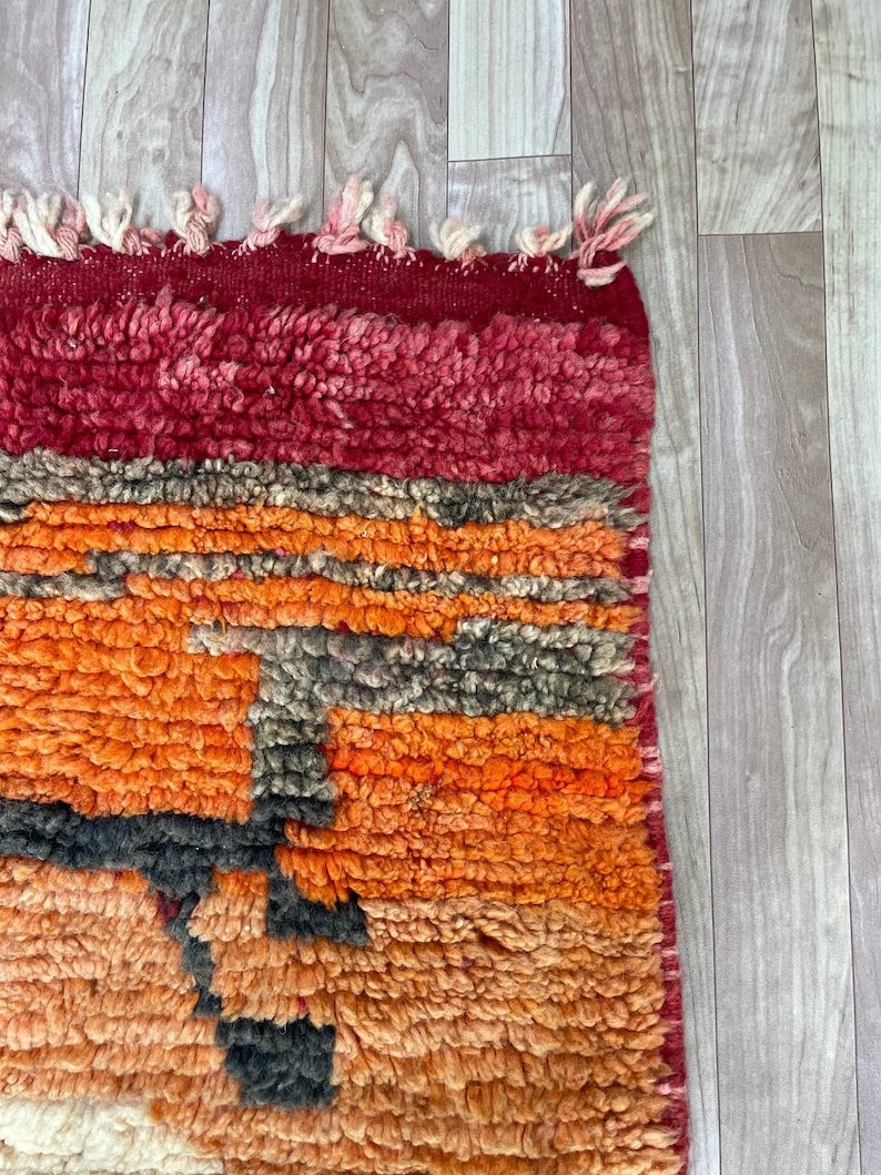 Puede incluir: Una alfombra marroqu&iacute; colorida con un patr&oacute;n geom&eacute;trico en tonos rojos, naranjas y negros. La alfombra tiene un borde de flecos y est&aacute; hecha de lana.