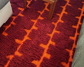 May include: A close-up of a shag rug with a geometric design. The rug is mainly a deep red, featuring a repeating pattern of orange shapes. The rug is on the floor near a wooden table, creating a warm, inviting atmosphere.