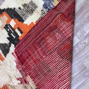 Puede incluir: Una alfombra tejida a mano con fondo blanco y patrones geom&eacute;tricos coloridos en tonos naranja, rojo, negro y gris. La alfombra est&aacute; doblada por la mitad, revelando el lado inferior rojo.