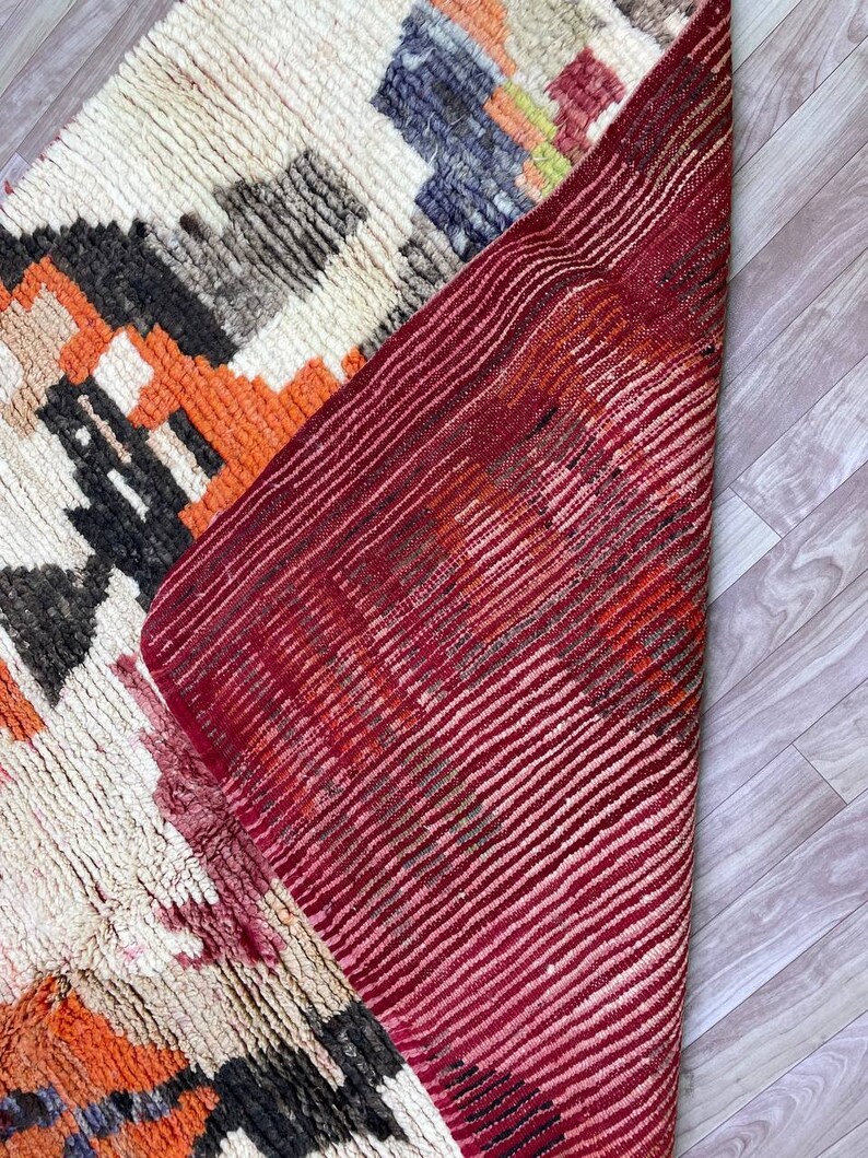 Puede incluir: Una alfombra tejida a mano con fondo blanco y patrones geom&eacute;tricos coloridos en tonos naranja, rojo, negro y gris. La alfombra est&aacute; doblada por la mitad, revelando el lado inferior rojo.