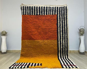 Pode incluir: Um tapete tecido à mão com um design geométrico em tons de laranja, amarelo, preto e branco. O tapete tem um pelo espesso e texturizado e é feito com fibras naturais.
