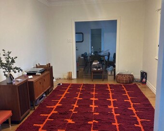 May include: A large, rectangular rug with a deep red background and a repeating orange geometric pattern. The rug is placed on a wooden floor in a room with wooden furniture. A black cat rests on the rug.