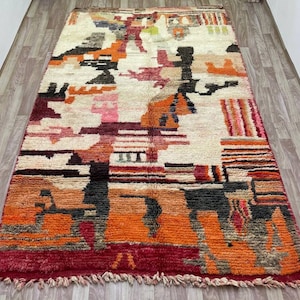 Puede incluir: Una alfombra abstracta colorida con un patr&oacute;n geom&eacute;trico en tonos de rojo, naranja, marr&oacute;n y blanco. La alfombra tiene un borde de flecos y est&aacute; hecha de lana.