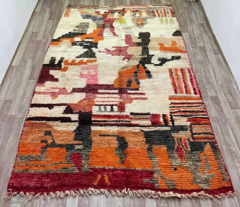 Puede incluir: Una alfombra abstracta colorida con un patr&oacute;n geom&eacute;trico en tonos de rojo, naranja, marr&oacute;n y blanco. La alfombra tiene un borde de flecos y est&aacute; hecha de lana.