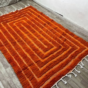 K&ouml;nnte beinhalten: Ein rechteckiger Teppich mit geometrischem Muster in Orange- und Oranget&ouml;nen. Das Design zeigt konzentrische Quadrate. Der Teppich hat eine strukturierte, gewebte Optik und wei&szlig;e Fransen an den Kanten. Er liegt auf einem Holzboden.