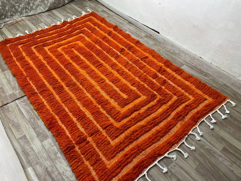 K&ouml;nnte beinhalten: Ein rechteckiger Teppich mit geometrischem Muster in Orange- und Oranget&ouml;nen. Das Design zeigt konzentrische Quadrate. Der Teppich hat eine strukturierte, gewebte Optik und wei&szlig;e Fransen an den Kanten. Er liegt auf einem Holzboden.
