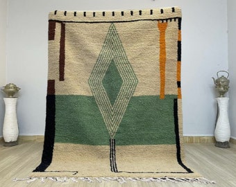 May include: A hand-woven rug with a geometric design in shades of beige, green, orange, and black. The rug features a large diamond shape in the center with a smaller diamond inside.