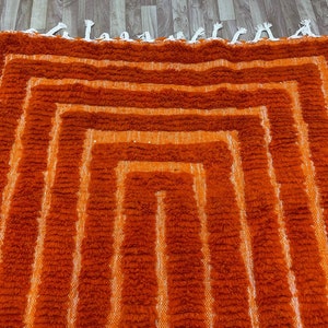 K&ouml;nnte beinhalten: Ein geometrischer Teppich in Orange- und Wei&szlig;t&ouml;nen mit einem strukturierten, zotteligen Aussehen. Der Teppich hat eine Reihe konzentrischer, rechteckiger Formen, die einen geschichteten Effekt erzeugen. Die Kanten sind mit wei&szlig;en Quasten versehen. Dieser Teppich ist f&uuml;r die Inneneinrichtung geeignet.