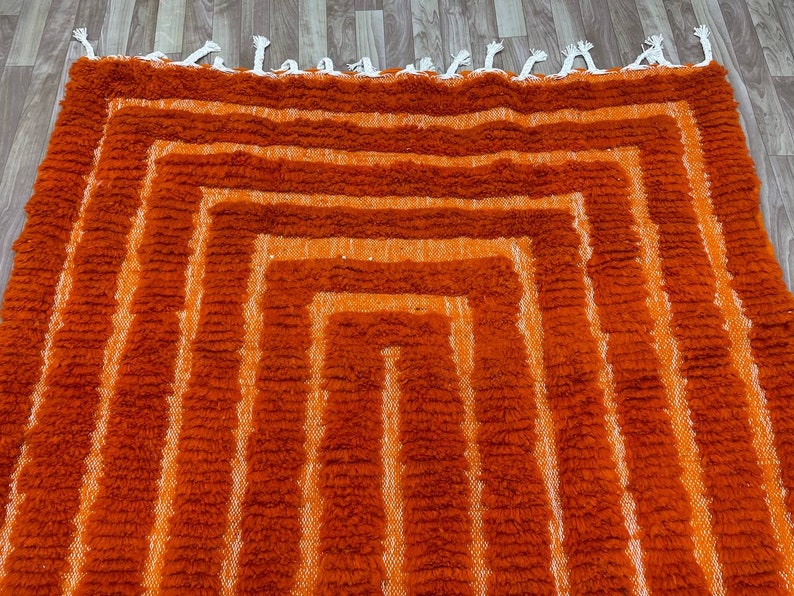 K&ouml;nnte beinhalten: Ein geometrischer Teppich in Orange- und Wei&szlig;t&ouml;nen mit einem strukturierten, zotteligen Aussehen. Der Teppich hat eine Reihe konzentrischer, rechteckiger Formen, die einen geschichteten Effekt erzeugen. Die Kanten sind mit wei&szlig;en Quasten versehen. Dieser Teppich ist f&uuml;r die Inneneinrichtung geeignet.