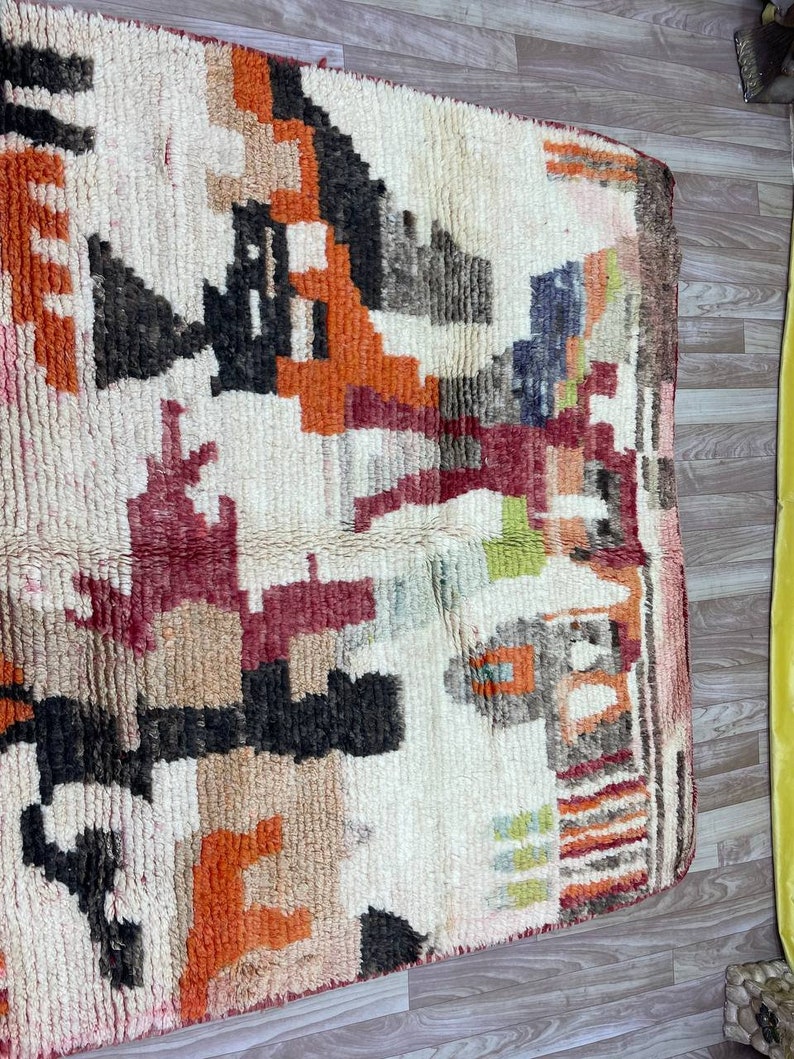 Puede incluir: Una alfombra vintage tejida a mano con un patr&oacute;n geom&eacute;trico en tonos de rojo, naranja, marr&oacute;n y blanco. La alfombra tiene un aspecto desgastado y descolorido, lo que a&ntilde;ade a su car&aacute;cter &uacute;nico.
