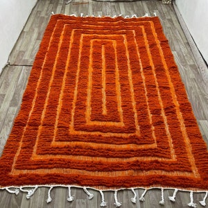 K&ouml;nnte beinhalten: Ein rechteckiger, orangefarbener Teppich mit geometrischem Design. Der Teppich weist eine Reihe konzentrischer Quadrate in verschiedenen Oranget&ouml;nen auf, die einen strukturierten, geschichteten Effekt erzeugen. Wei&szlig;e Quasten s&auml;umen die R&auml;nder.