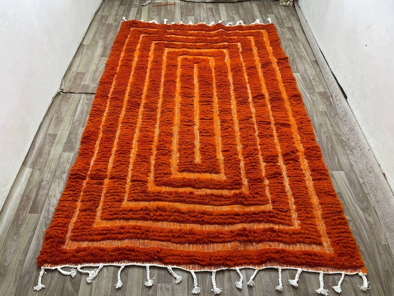 K&ouml;nnte beinhalten: Ein rechteckiger, orangefarbener Teppich mit geometrischem Design. Der Teppich weist eine Reihe konzentrischer Quadrate in verschiedenen Oranget&ouml;nen auf, die einen strukturierten, geschichteten Effekt erzeugen. Wei&szlig;e Quasten s&auml;umen die R&auml;nder.