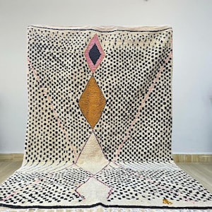 Puede incluir: Una alfombra marroquí blanca con un patrón geométrico en negro, rosa y naranja. La alfombra tiene una forma de diamante en el centro con un diamante más pequeño en el interior. La alfombra está hecha de lana y tiene flecos en los bordes.