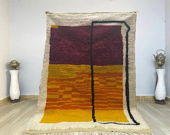 Puede incluir: Una alfombra de lana tejida a mano con un diseño geométrico en tonos burdeos, naranja y amarillo. La alfombra tiene un borde blanco y un contorno negro alrededor de las formas geométricas.