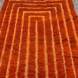 K&ouml;nnte beinhalten: Ein rechteckiger Teppich in Orange- und Wei&szlig;t&ouml;nen mit geometrischem Muster. Das Muster des Teppichs besteht aus konzentrischen Rechtecken, wobei wei&szlig;e Streifen die orangefarbenen Abschnitte trennen. Der Teppich hat ein strukturiertes, zotteliges Aussehen.