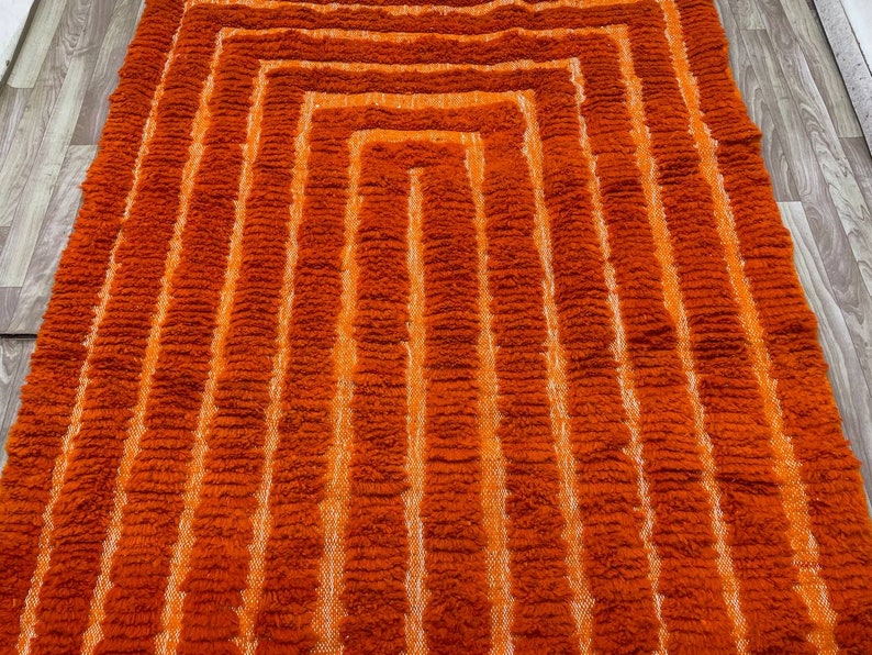 K&ouml;nnte beinhalten: Ein rechteckiger Teppich in Orange- und Wei&szlig;t&ouml;nen mit geometrischem Muster. Das Muster des Teppichs besteht aus konzentrischen Rechtecken, wobei wei&szlig;e Streifen die orangefarbenen Abschnitte trennen. Der Teppich hat ein strukturiertes, zotteliges Aussehen.