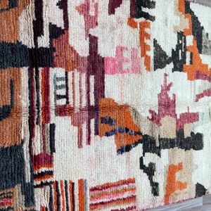 Puede incluir: Una alfombra tejida a mano con un patr&oacute;n geom&eacute;trico en tonos de naranja, rojo, rosa, negro y blanco. La alfombra tiene un fondo de color crema y una superficie texturizada y esponjosa.