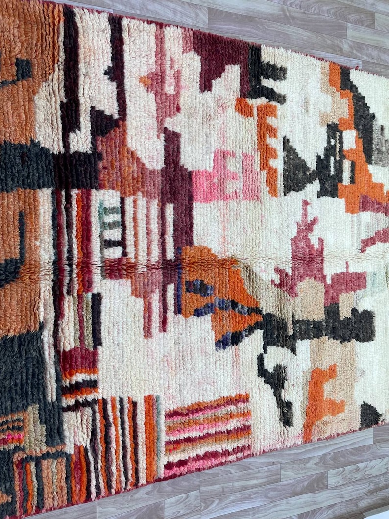 Puede incluir: Una alfombra tejida a mano con un patr&oacute;n geom&eacute;trico en tonos de naranja, rojo, rosa, negro y blanco. La alfombra tiene un fondo de color crema y una superficie texturizada y esponjosa.
