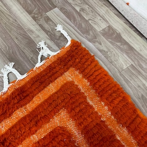 K&ouml;nnte beinhalten: Nahaufnahme eines lebendigen orange-wei&szlig; geometrisch gemusterten Teppichs. Der Teppich hat einen strukturierten, zotteligen Flor mit einem sich wiederholenden Design aus orangefarbenen Streifen und wei&szlig;en Akzenten. Wei&szlig;e Quasten schm&uuml;cken den Rand des Teppichs.