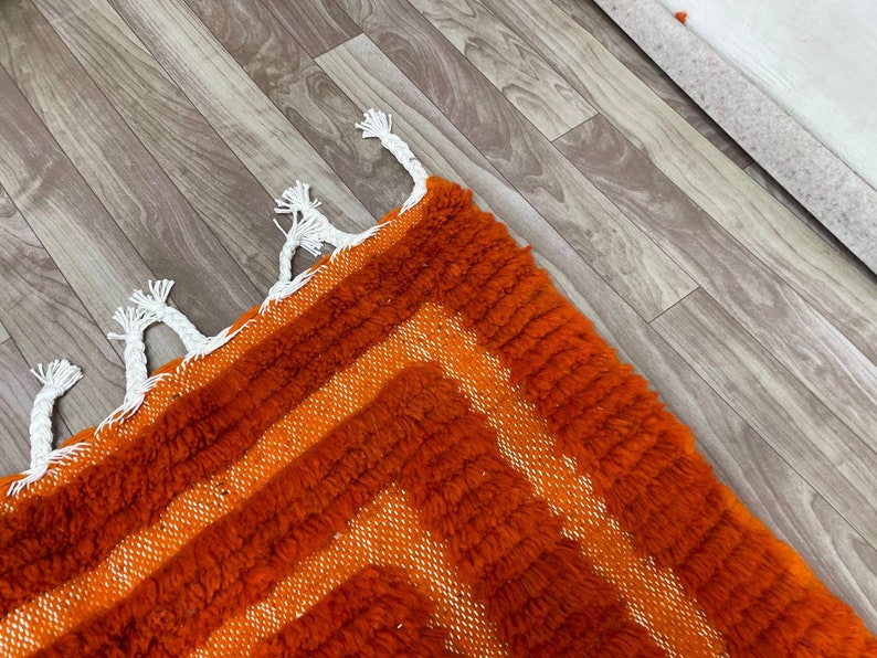 K&ouml;nnte beinhalten: Nahaufnahme eines lebendigen orange-wei&szlig; geometrisch gemusterten Teppichs. Der Teppich hat einen strukturierten, zotteligen Flor mit einem sich wiederholenden Design aus orangefarbenen Streifen und wei&szlig;en Akzenten. Wei&szlig;e Quasten schm&uuml;cken den Rand des Teppichs.