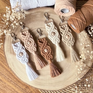 Könnte beinhalten: Fünf handgefertigte Makramee-Schlüsselanhänger mit Quasten. Jeder Schlüsselanhänger hat ein anderes Design und eine andere Farbkombination, darunter Weiß, Braun und Beige. Die Schlüsselanhänger sind aus Naturfasern gefertigt und haben einen rustikalen, bohemischen Look.