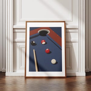 May include: A framed print depicting a pool table illustration. The table has a blue playing surface, red rails, and a pocket. Several billiard balls, including red, white, and black, are scattered across the table, along with a cue stick.