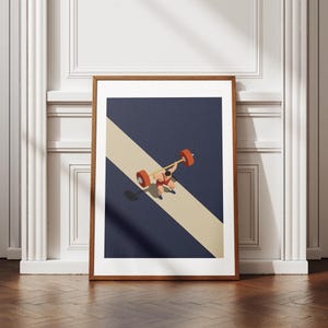 Op de afbeelding: Een ingelijste print met een gewichtheffer in het rood die een halter met oranje gewichten optilt. De achtergrond is marineblauw met een diagonale beige streep. De print wordt op een witte muur weergegeven.