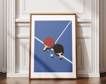 Op de afbeelding: Een ingelijste print met een gestileerde pingpongscène. Een rode en een zwarte bat liggen op een blauw oppervlak met witte lijnen, samen met een witte bal. De print zit in een bruine houten lijst, ideaal voor sportliefhebbers.