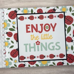 May include: A decorative notebook with a black spine and a cover featuring a ladybug and floral pattern. The cover has the words "ENJOY the little THINGS" in red, yellow, and green. The edge is decorated with white pom-poms.