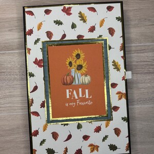 Può includere: Un diario rettangolare a tema autunnale. Ha uno sfondo bianco ornato di foglie autunnali colorate. Il centro presenta un rettangolo arancione con girasoli, zucche e la scritta "FALL is my Favorite". Il diario ha un bordo nero.