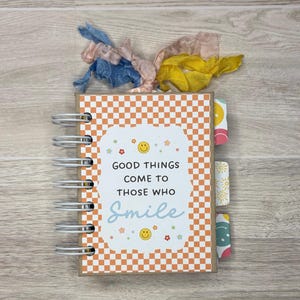 May include: A spiral-bound notebook with an orange and white checkered cover. The cover features the text "Good Things Come to Those Who Smile" with smiley face and flower graphics. The notebook has colorful ribbon accents.