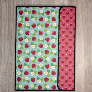 May include: A rectangular folder featuring a light blue background with a repeating pattern of red strawberries, green leaves, and white flowers, contrasted by a vertical side panel in dark pink with a repeating pattern of small red strawberries.