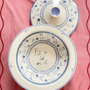 Op de afbeelding: Een witte keramische kom met een blauw bloemenpatroon en een bijpassend deksel. De kom heeft de woorden "Put a lid on it." aan de binnenkant.