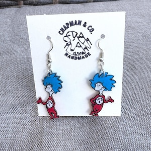 Puede incluir: Un par de pendientes colgantes con los personajes Thing 1 y Thing 2 del libro del Dr. Seuss. Los pendientes son rojos, azules y blancos, con ganchos plateados. Se muestran en una tarjeta blanca con el texto "CHAPMAN & CO. PLUM HANDMADE".
