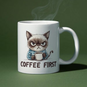 Può includere: Tazza in ceramica bianca con l'illustrazione di un gatto imbronciato che tiene una tazza di caffè. Il gatto indossa una camicia a quadri blu. La tazza ha la scritta "COFFEE FIRST" in lettere marroni. Vapore che sale dalla tazza su sfondo verde.