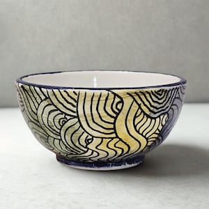 Puede incluir: Un cuenco de cerámica con interior blanco y borde y base azul oscuro. El exterior presenta un diseño pintado a mano de líneas negras onduladas sobre un fondo de tonos verdes y amarillos. El cuenco es una pieza decorativa.