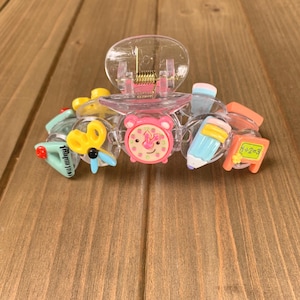 May include: Clear plastic hair claw clip decorated with school-themed charms. The charms include a pink clock, a pencil, a chalkboard with a math problem, a plane, and a pair of scissors. The clip is set against a wooden background.