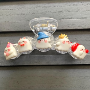 Puede incluir: Pinza para el pelo de plástico transparente con cinco dijes blancos con forma de fantasma, cada uno con accesorios únicos como sombreros y lazos. Los dijes tienen detalles en rosa, rojo y azul. La pinza está diseñada para sujetar el cabello.