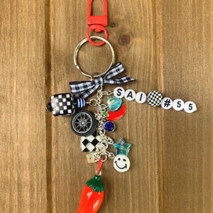 May include: A decorative keychain with a red clip, silver ring, and various charms. Charms include a checkered flag, a car wheel, a red chili pepper, and a smiley face. The keychain also features the letters "SAI" and the number "55" on white beads.