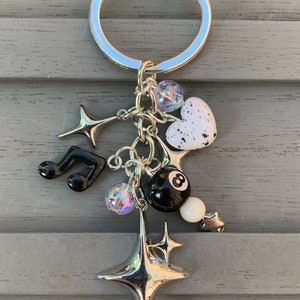 Puede incluir: Un llavero plateado con una variedad de dijes, que incluyen un corazón blanco y negro, una bola de billar blanca y negra, una estrella plateada y una nota musical negra.