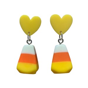 May include: A pair of yellow heart dangle earrings with a candy corn charm. The candy corn charm is yellow, orange, and white.