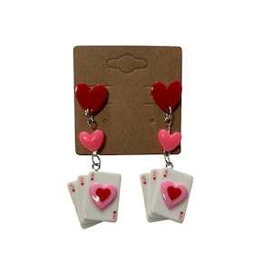 May include: A pair of dangle earrings featuring a red heart, a pink heart, and a stack of playing cards with a red heart on the face card. The cards are the Ace, 2, 4, and 5 of hearts.