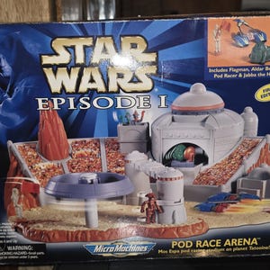 Op de afbeelding: Star Wars Episode I Micro Machines Pod Race Arena speelset. De doos heeft een blauwe achtergrond met een personage met een lichtzwaard. De set bevat een stadion, podracers en figuren. Eerste editie.