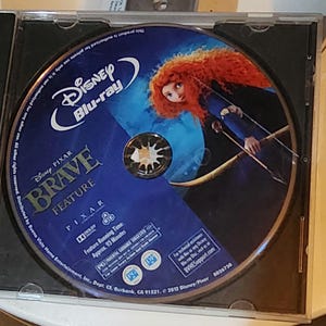 Puede incluir: Un disco Blu-ray azul y dorado con la pel&iacute;cula Brave de Disney Pixar. El disco presenta un primer plano de Merida, el personaje principal, con su cabello rojo y arco y flecha. El texto en el disco dice "Disney Pixar Brave Feature Pixar Blu-ray".
